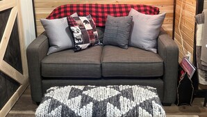 Ein gemütliches Zimmer mit einer Holzwand, einem grauen Sofa mit kariertem Überwurf und einem Scrabble-Spiel auf einem kleinen Tisch.