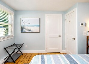 Ein Schlafzimmer mit Bett, Tür, einem Fenster mit Jalousien, einem Wandbild und einem Schuhregal.