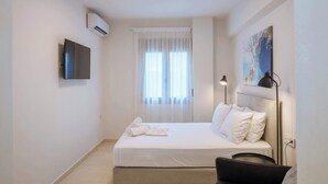 Habitación de hotel con cama, televisión de pantalla plana, aire acondicionado, ventana con cortinas translúcidas y un cuadro en la pared.
