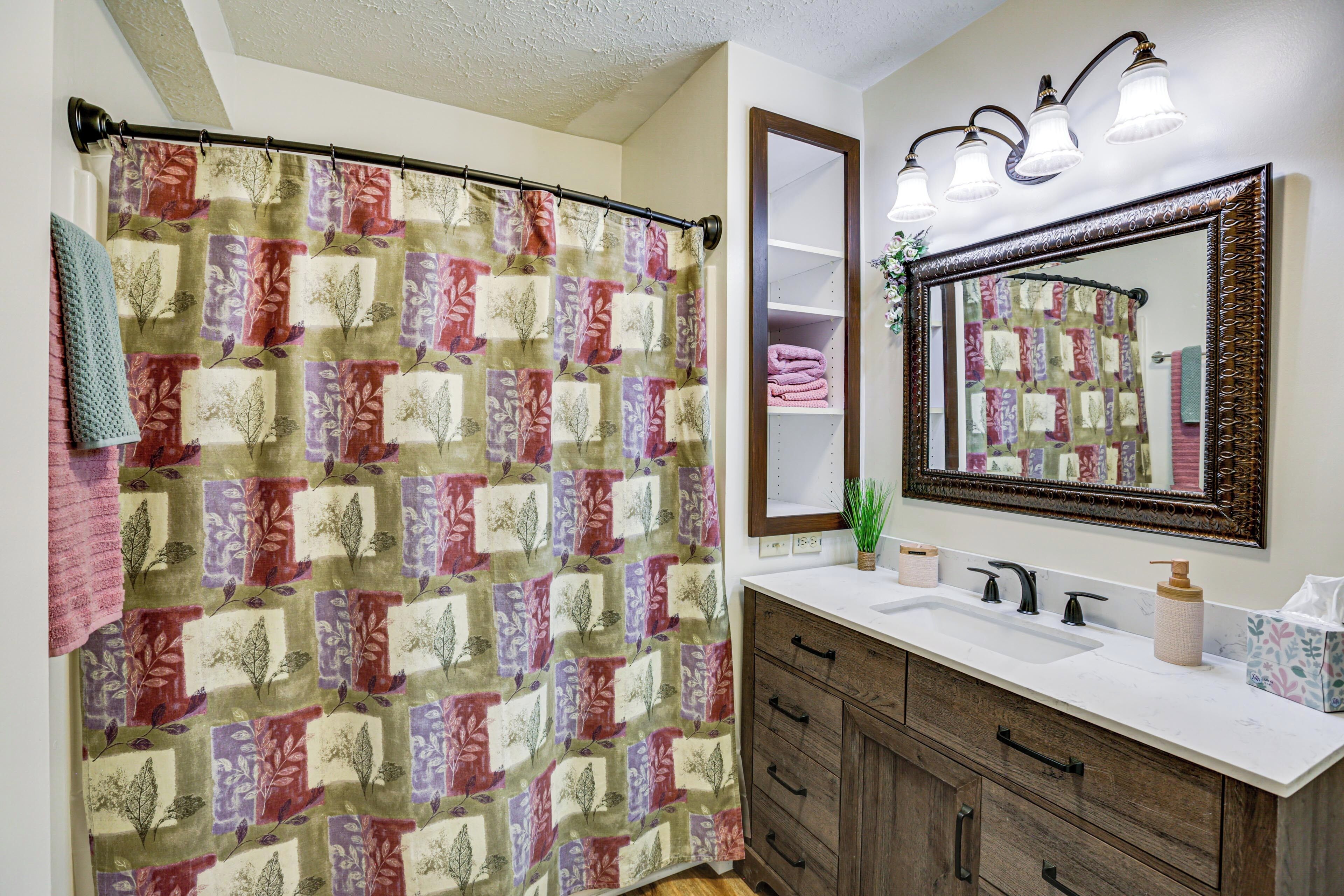 A bathroom with a quilted shower curtain, a large mirror, a sink with two faucets, and wooden cabinets.