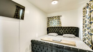 A bedroom with a tufted headboard, a flat-screen TV mounted on the wall, a bed with a patterned bedspread, and floral curtains. - Walk to Shops, Parks & Restaurants: Teaneck Escape (Teaneck)