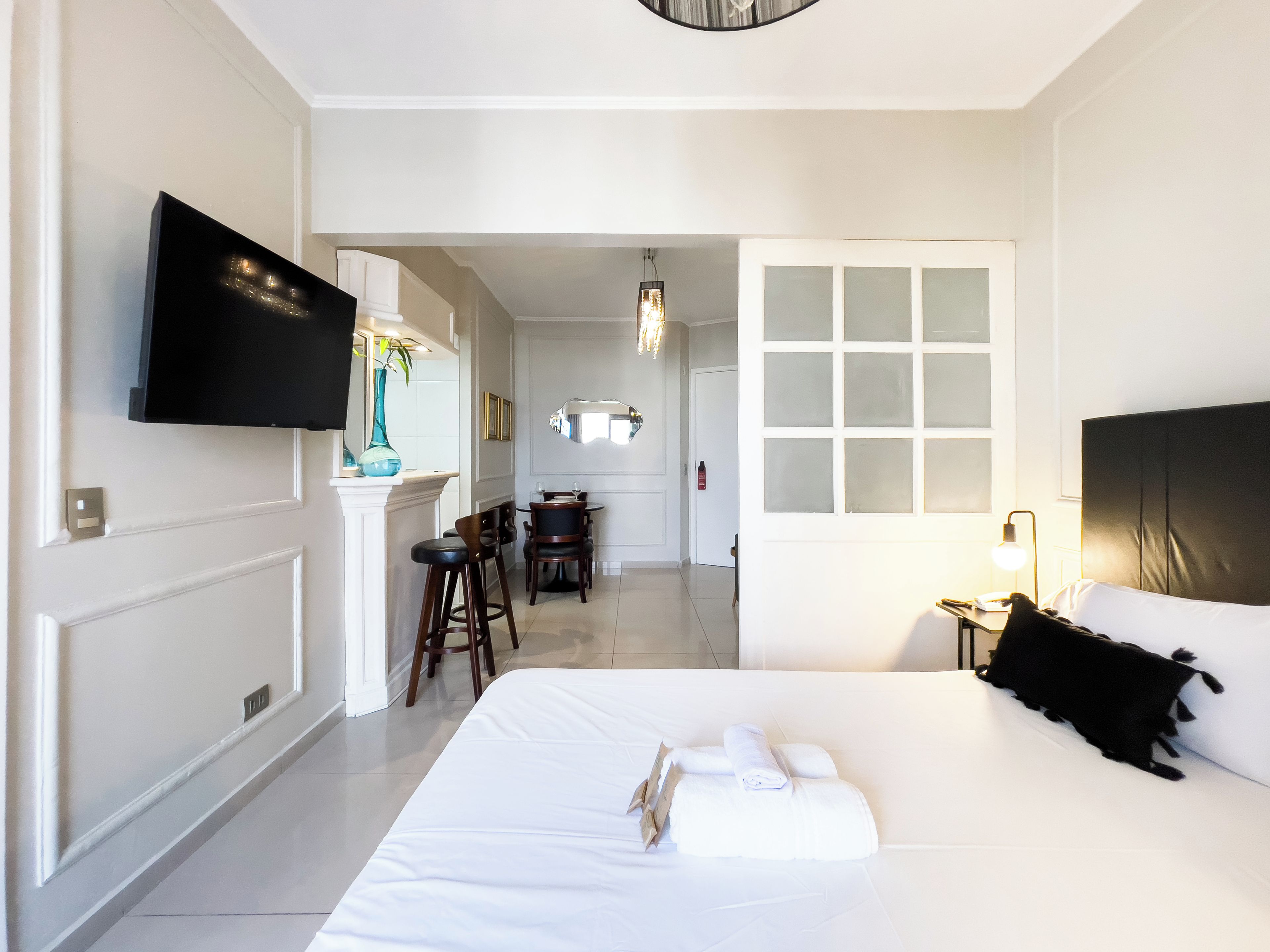 A hotel room with a bed, a TV mounted on the wall, a dining area with chairs and a table, and a hallway leading to another room.