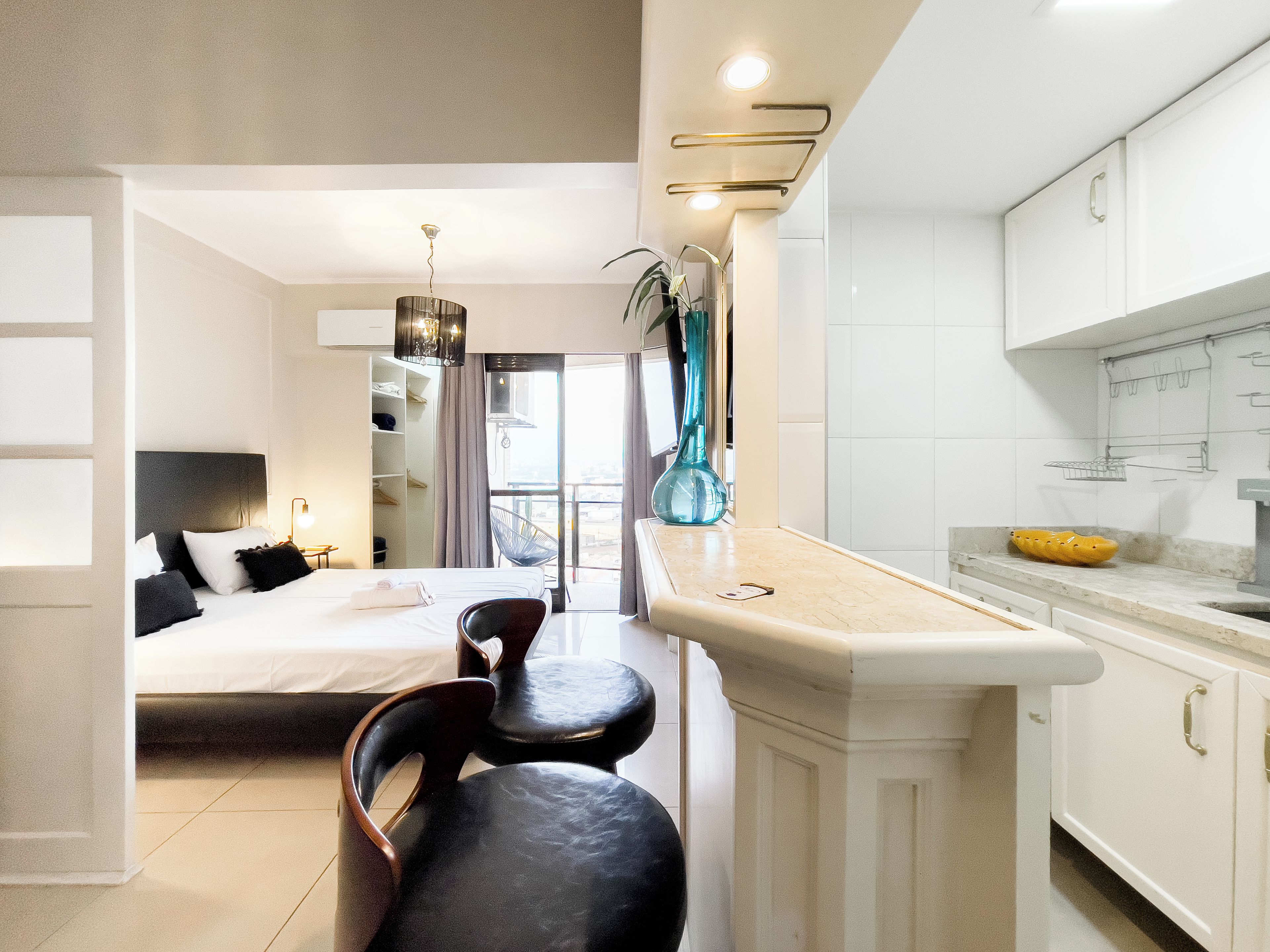 A modern hotel room with a bed, seating area, kitchenette, and a view through a glass door.