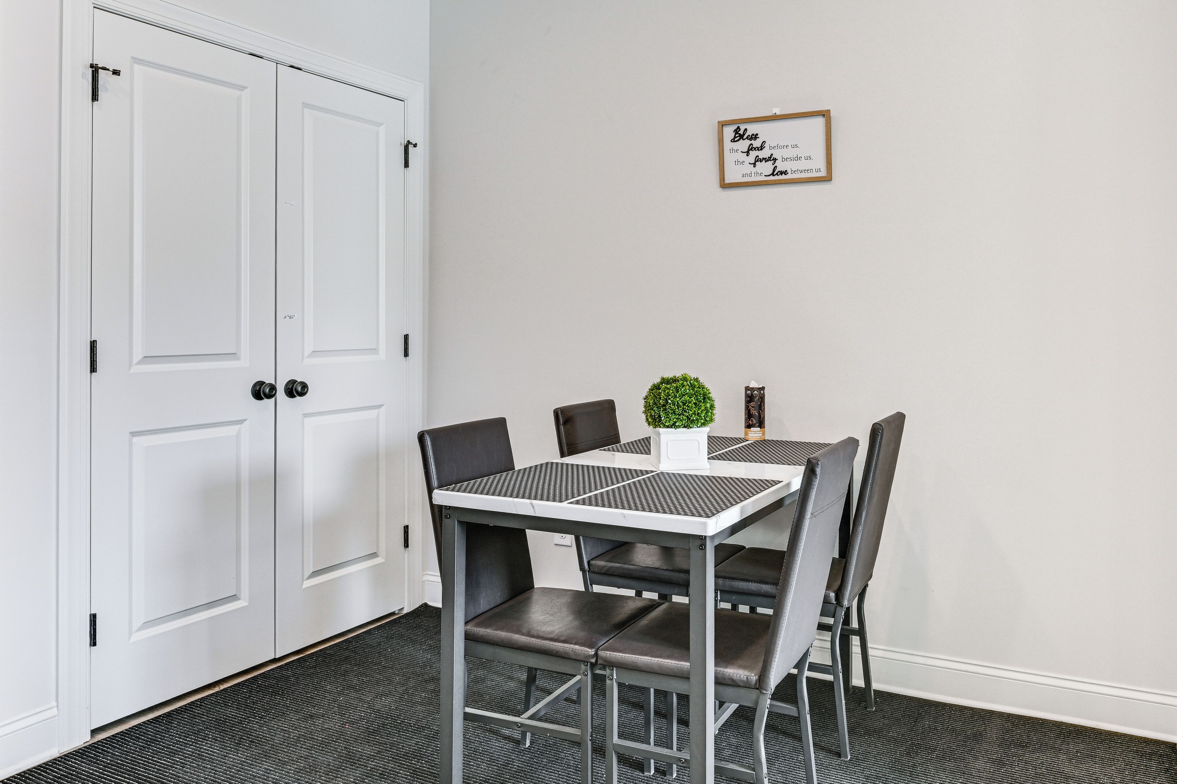 A room with a white door, a table with four chairs, a small plant on the table, and a framed quote on the wall.