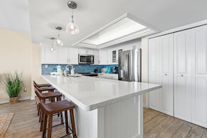 A modern kitchen with a central island, white cabinetry, stainless steel appliances, and wooden bar stools. - Updated Corner 3BR w/ View: Suntide III - Unit 710 (South Padre Island)