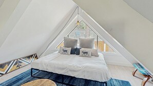 Une chambre avec un plafond en pente, un lit avec une literie blanche et des coussins décoratifs, un tapis bleu et un banc.