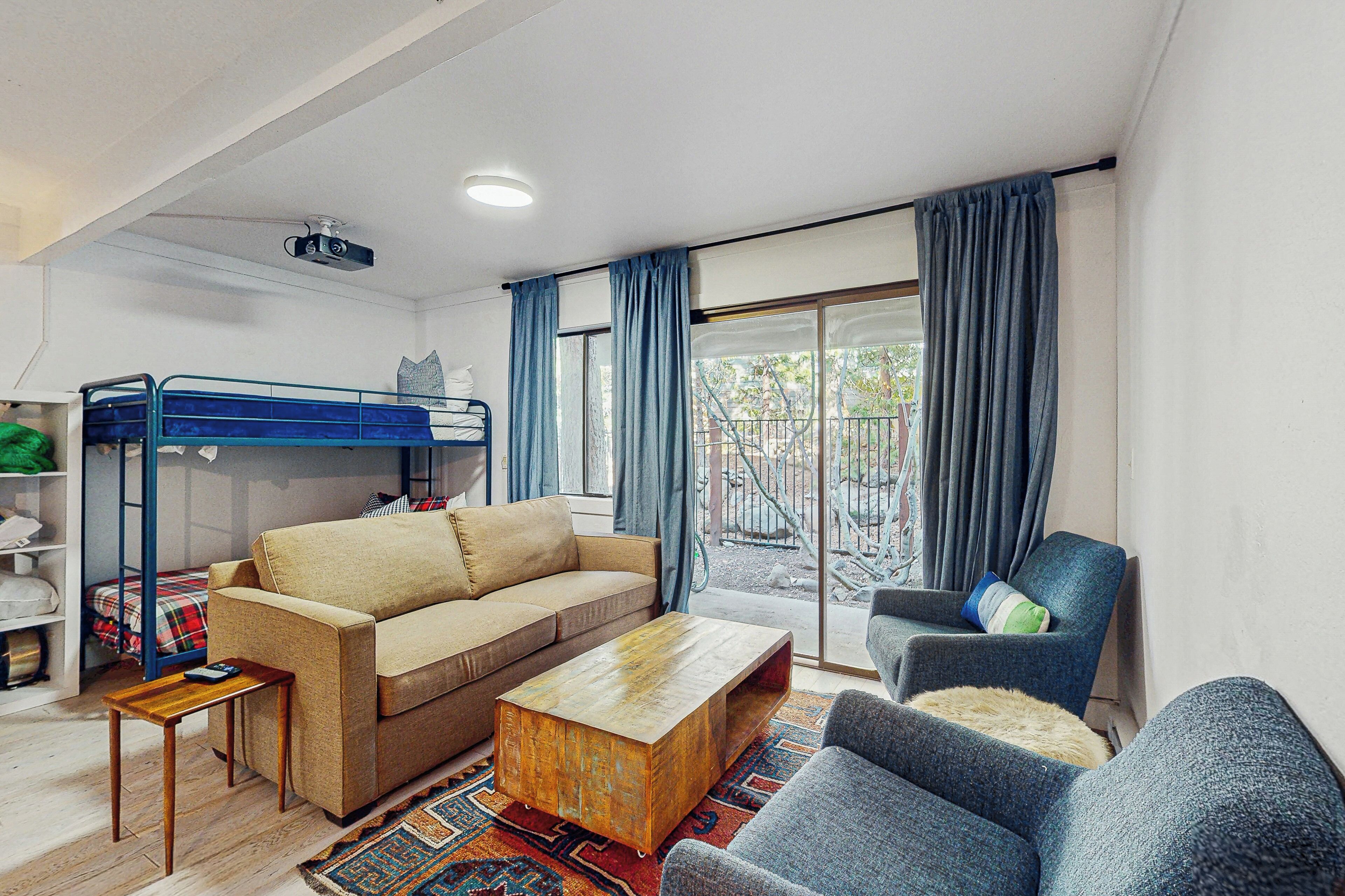 A compact living space with a sofa, a coffee table, a chair, a bunk bed, and a sliding glass door leading to an outdoor area.