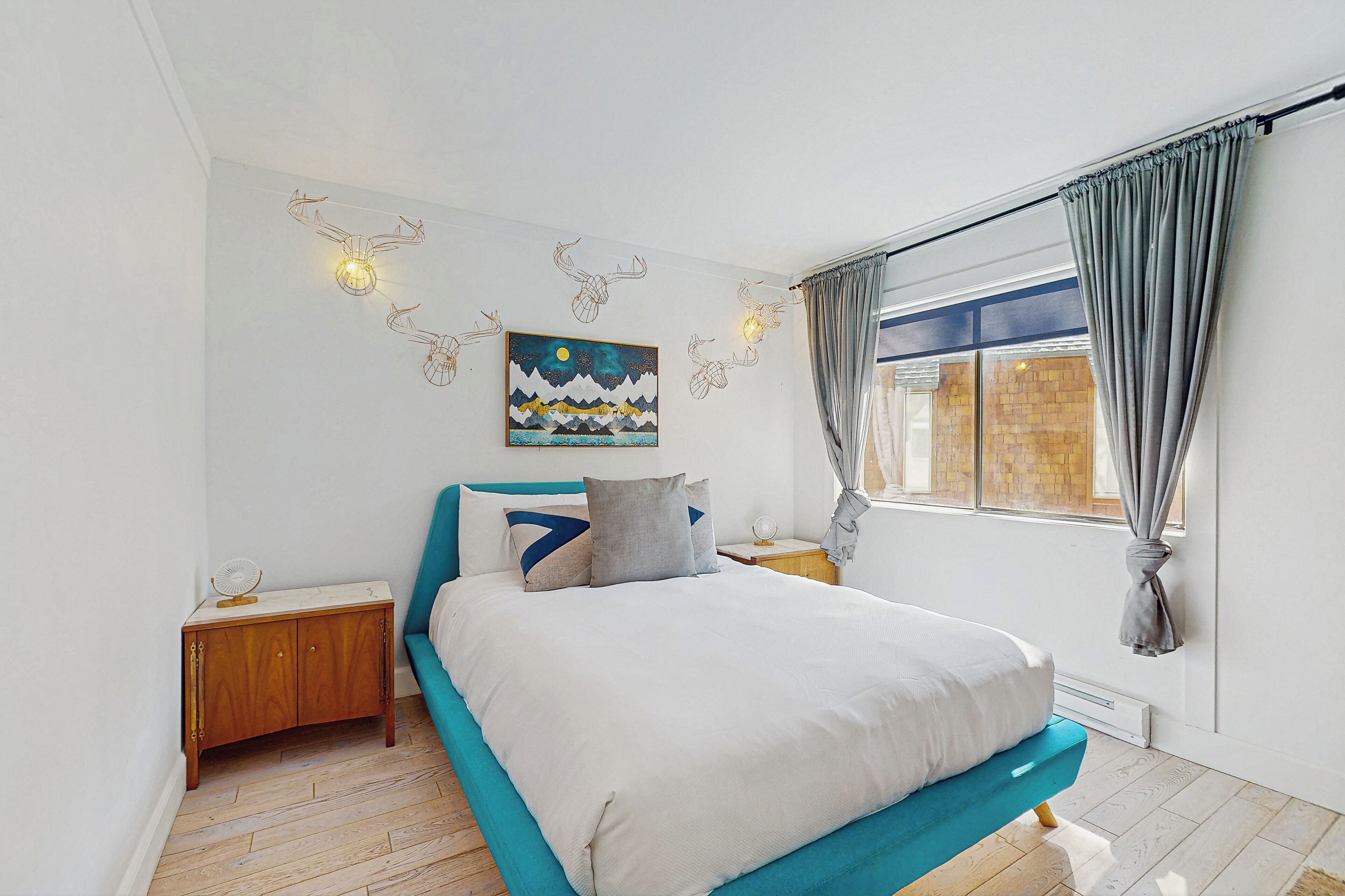 Ein Schlafzimmer mit einem Bett, einem Nachttisch, einem Wandventilator, einem Gemälde und einem Fenster mit Vorhängen.