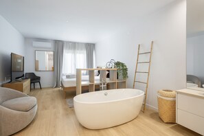 Ein modernes, helles Schlafzimmer mit einem großen Fenster, einem integrierten Schreibtisch, einem bequemen Sessel und einer großzügigen Badewanne.