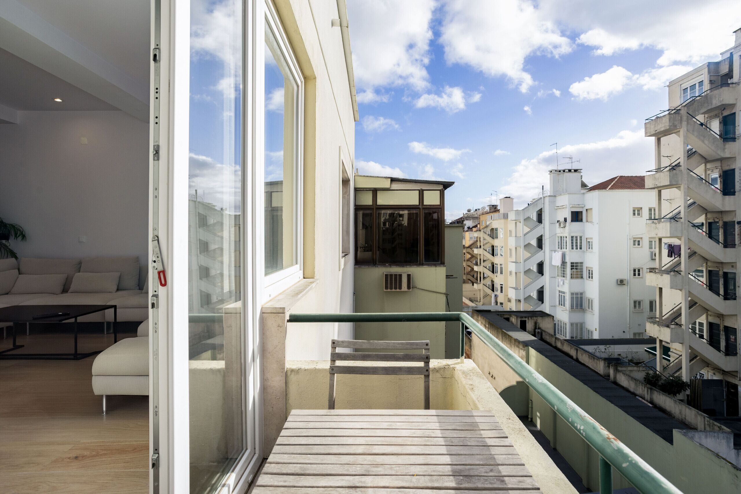 A balcony with a wooden table and a chair, overlooking a cityscape with multiple buildings.