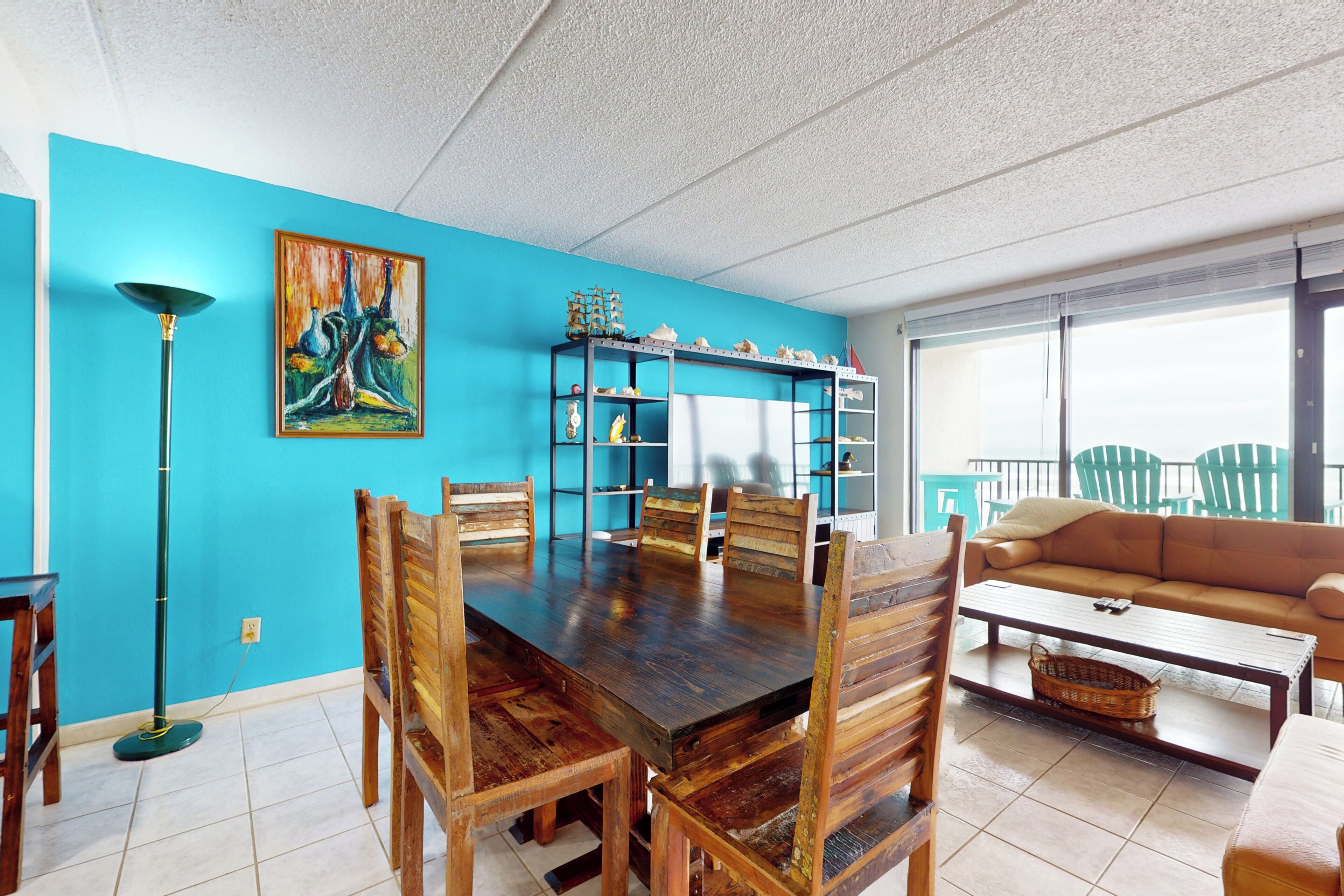Un coin repas avec une table et des chaises en bois, un mur bleu orné d’un tableau, et un canapé donnant sur l’extérieur.