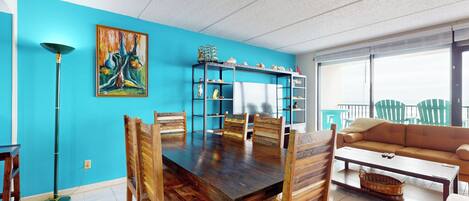 Un coin repas avec une table et des chaises en bois, un mur bleu orné d’un tableau, et un canapé donnant sur l’extérieur.