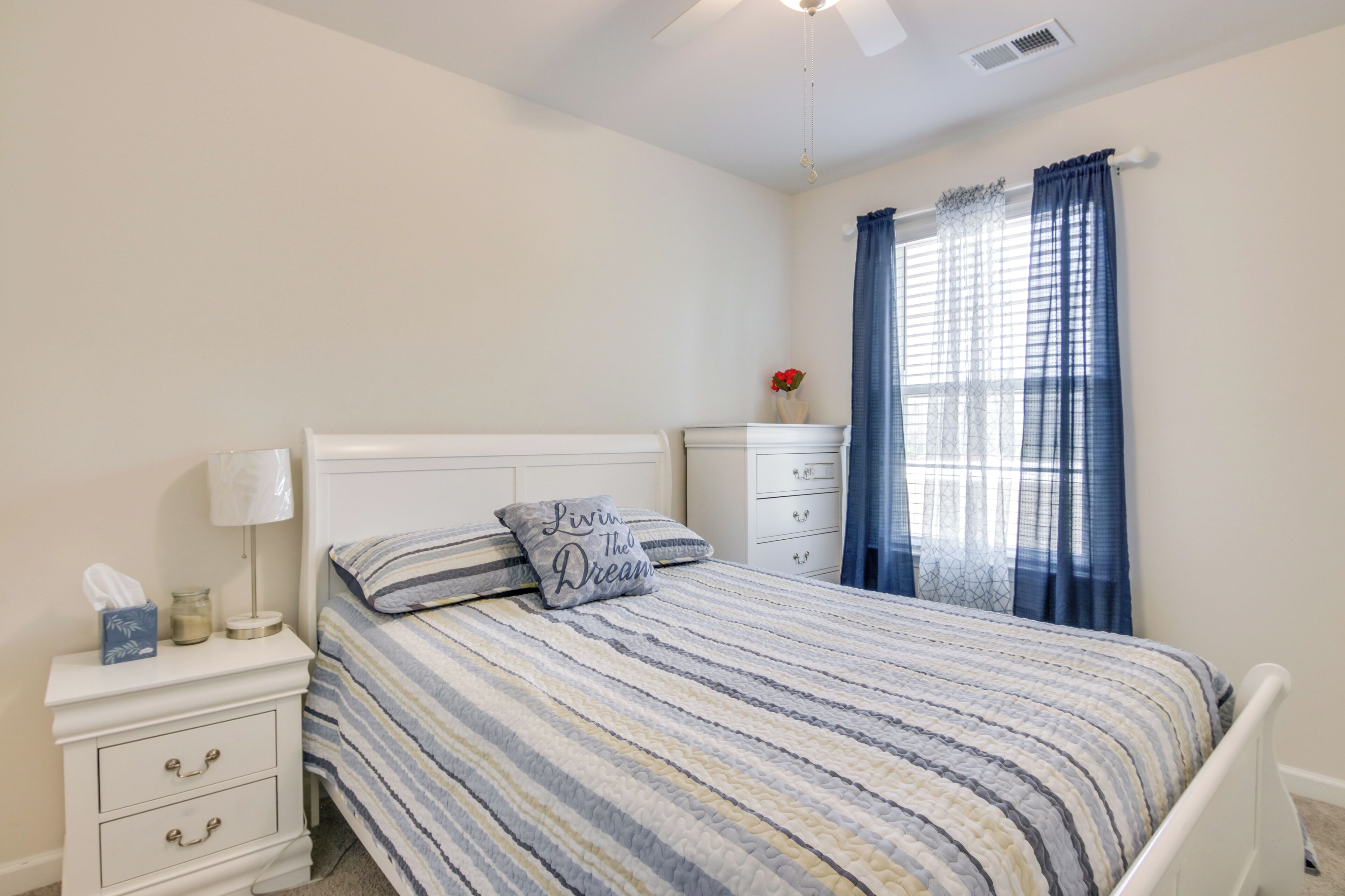 Une chambre à coucher comprenant un lit, une table de chevet, une lampe, une fenêtre avec des rideaux et un ventilateur de plafond.