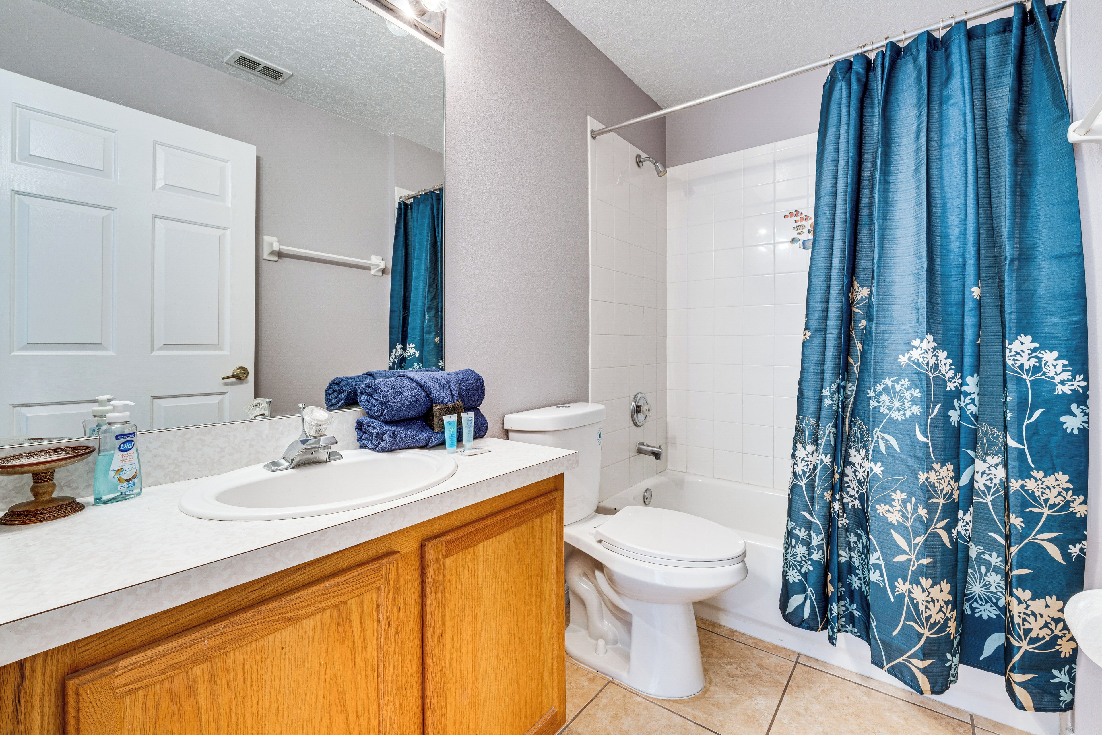 Un baño con lavamanos blanco, armarios de madera, inodoro y ducha con cortina azul estampada de flores.
