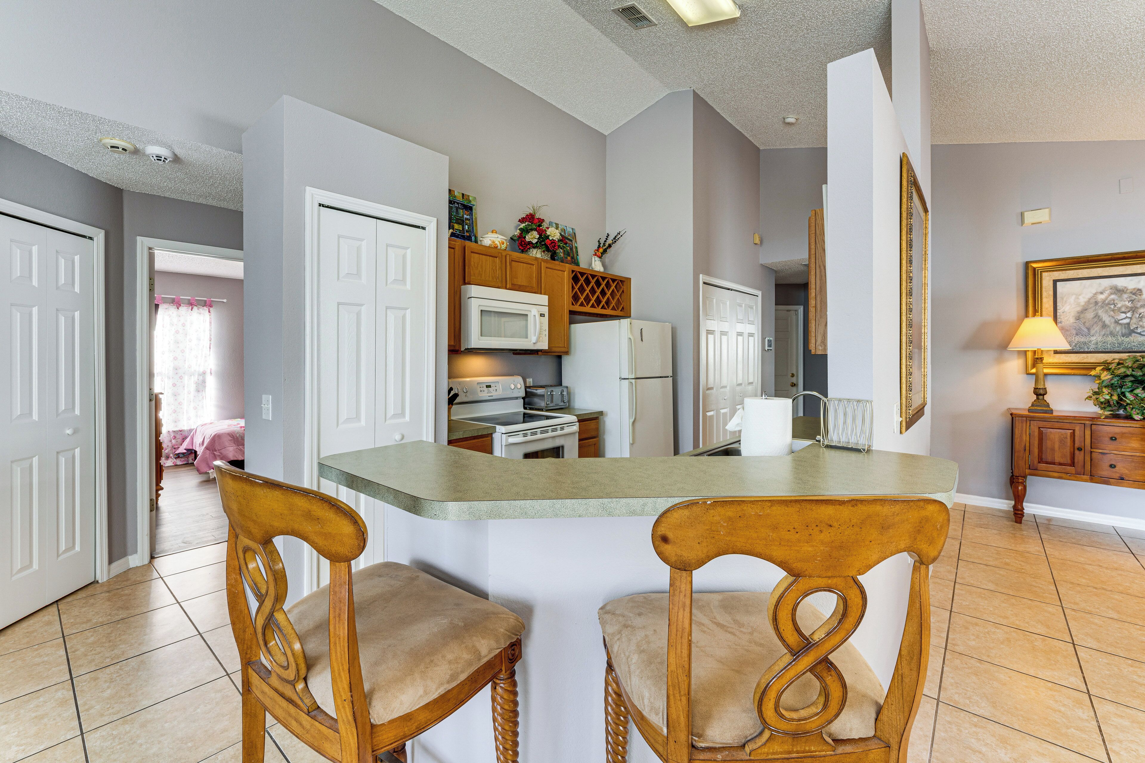 A kitchen with white cabinets, a microwave, and a refrigerator. There are two wooden chairs with cushioned seats.