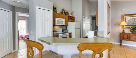 A kitchen with white cabinets, a microwave, and a refrigerator. There are two wooden chairs with cushioned seats.