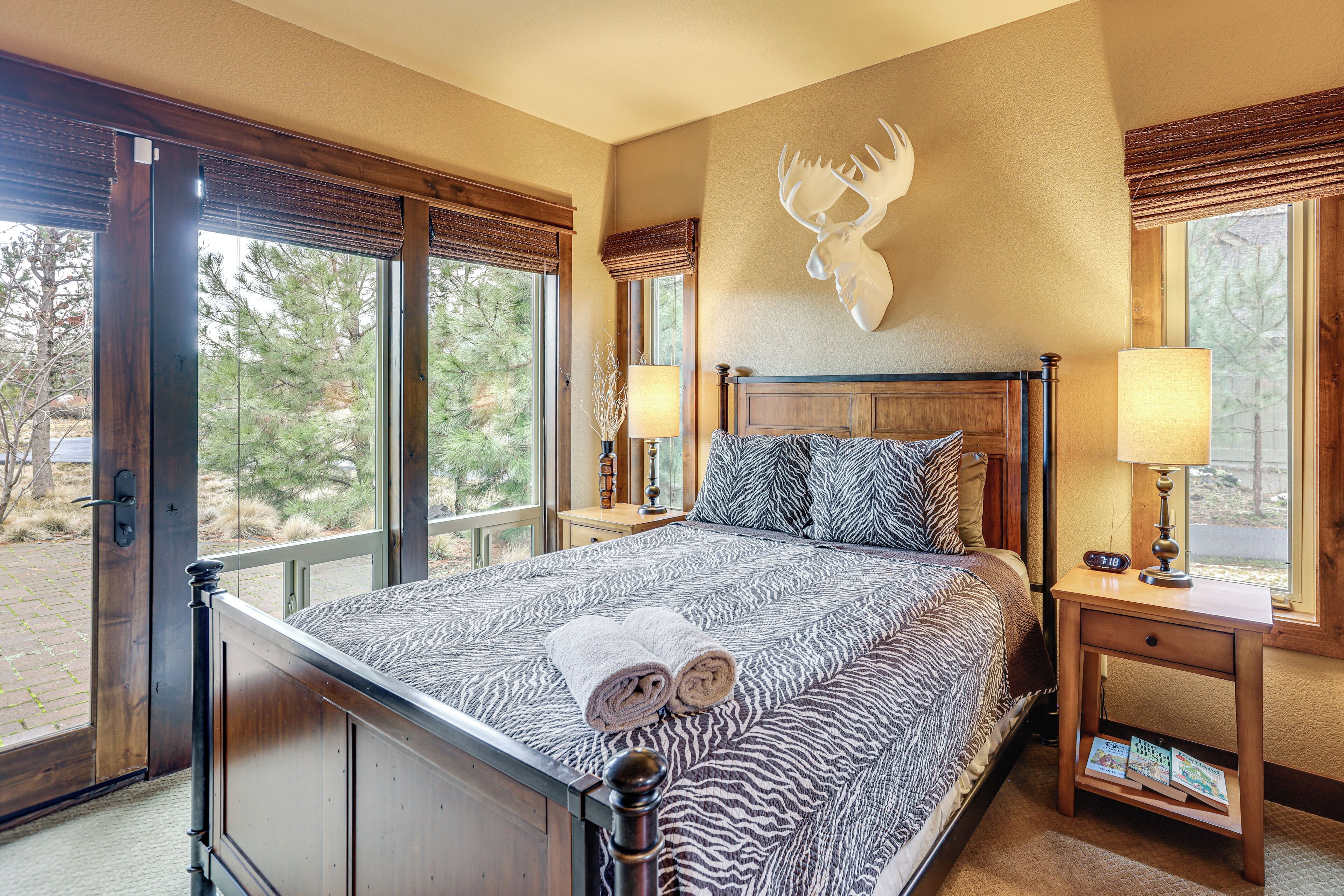 A bedroom with a large bed, two bedside tables with lamps, a mounted moose head on the wall, and a window with a view of trees.