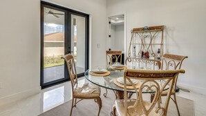 A dining area with a glass table and wicker chairs, a decorative metal shelf with glassware, and a view through a glass door to the outdoors. - Home Gym & Sauna! Luxe Lehigh Acres Family Home (Lehigh Acres)