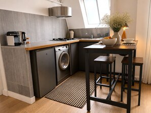 A compact kitchen with a washing machine, a coffee maker, and a dining area with stools. - L'appartement Haudriettes (Paris03)