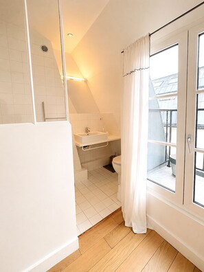 A bathroom with a shower, sink, and toilet, a window with curtains, and a view of the outside. - L'appartement Haudriettes (Paris03)