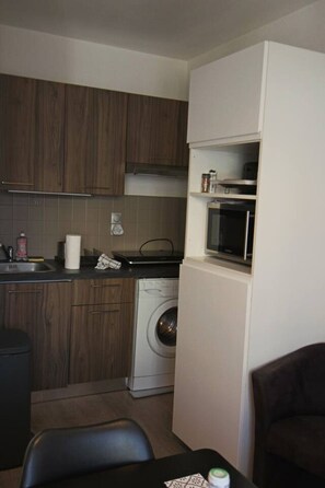 A compact kitchen with wooden cabinets, a washing machine, a microwave, and a small dining area with a table and chairs. - Studio 25 m², balcon en hyper centre-ville (Aix-les-Bains)