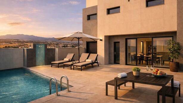 Un área de piscina moderna al aire libre con sillones de descanso, una mesa con frutas y vino, y vistas a la ciudad y las montañas.