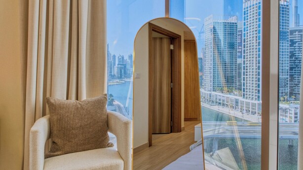 Una camera d'albergo moderna con una grande finestra che offre vista sulla città, un'area salotto accogliente con una poltrona imbottita e una porta in legno che conduce a un'altra stanza.
