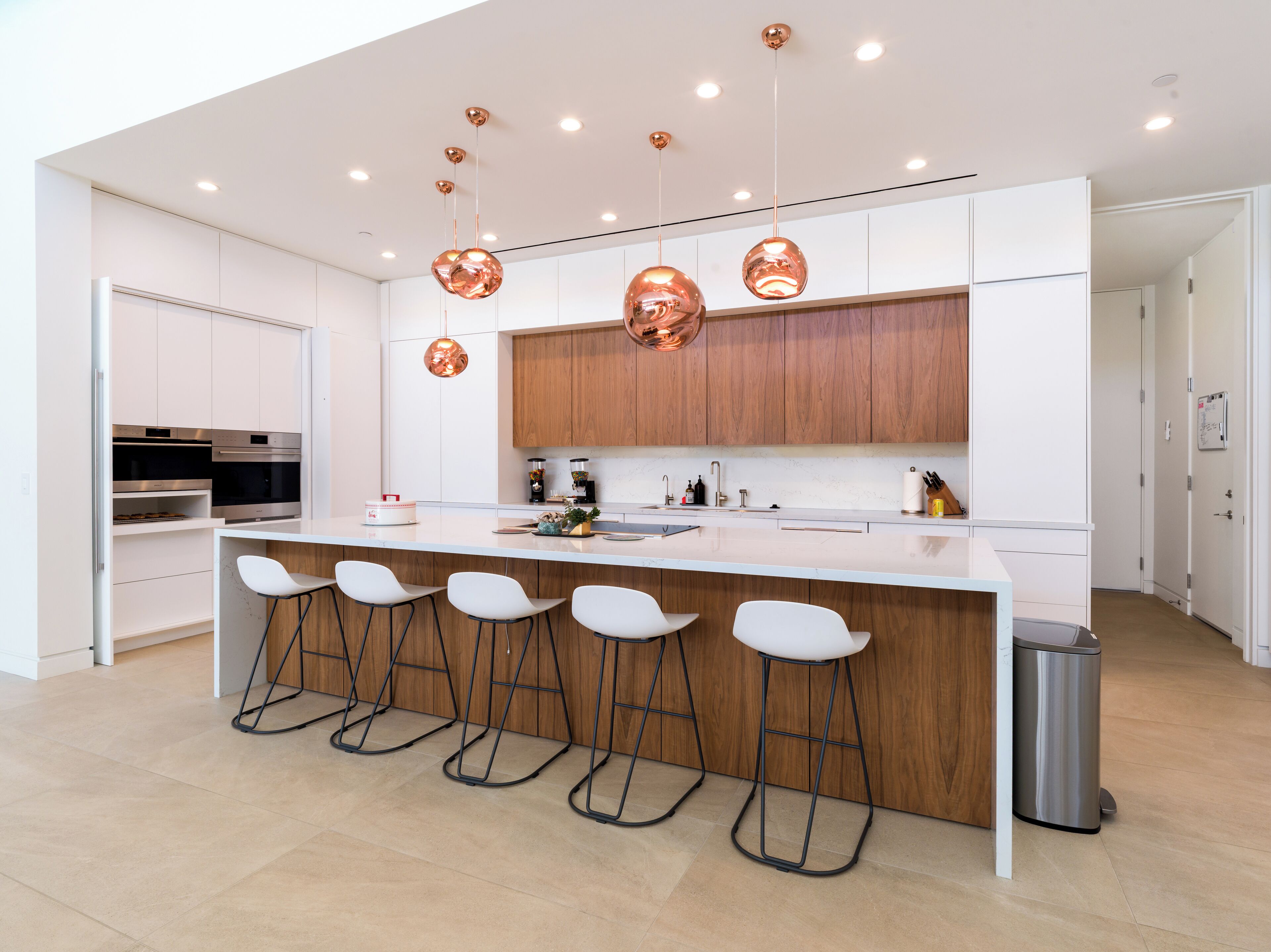 A modern kitchen with white cabinetry, a central island with bar stools, stainless steel appliances, and copper pendant lighting.