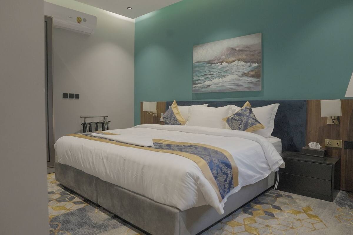 A modern hotel room with a large bed, a painting of a seascape, and a geometric patterned floor.