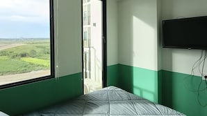 A bedroom with a large window, an air conditioner, a bed, and a mounted TV. - Apartemen Osaka Riverview by Linkara (Teluknaga)