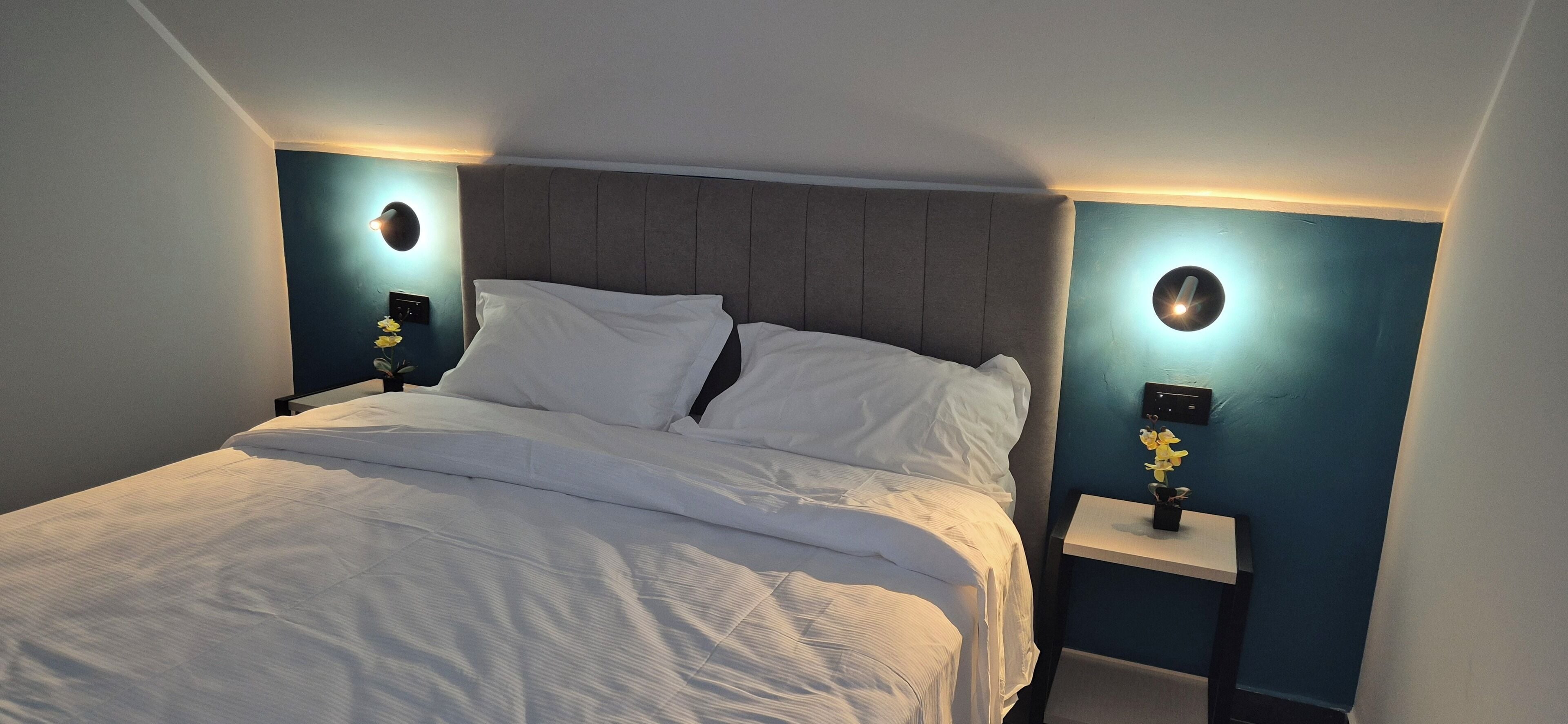 Una cama impecablemente arreglada con ropa de cama blanca, un cabecero tapizado en gris y dos mesitas de noche con lámparas y jarrones de flores.
