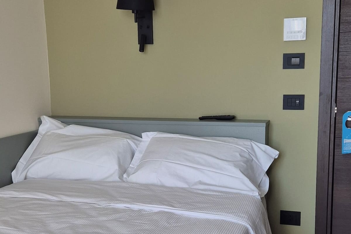 A neatly made bed with white linens in a room with a green wall, a black wall-mounted lamp, and a door with a blue handle.