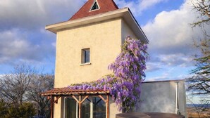 Un edificio a un piano con tetto rosso, una piccola finestra e un pergolato fiorito di fiori viola.
