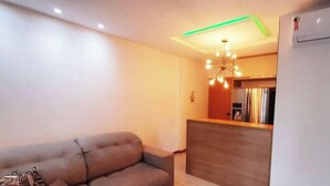Un salon moderne avec un canapé, une table basse en bois et un coin cuisine équipé d’un réfrigérateur et d’un luminaire suspendu.