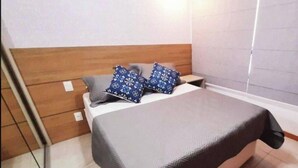 Un lit bien fait, avec une couvre-lit grise et deux coussins à motifs, une tête de lit en bois et un mur blanc avec une fenêtre.