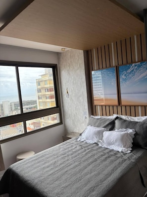 Une chambre avec une grande fenêtre, un lit recouvert d’une literie à motifs, une lampe fixée au mur et un tableau représentant une plage.