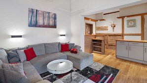 Una sala de estar moderna con un sofá seccional gris, una mesa redonda y una almohada roja. Un cuadro de un bosque está en la pared, y un reloj sobre la encimera de la cocina.