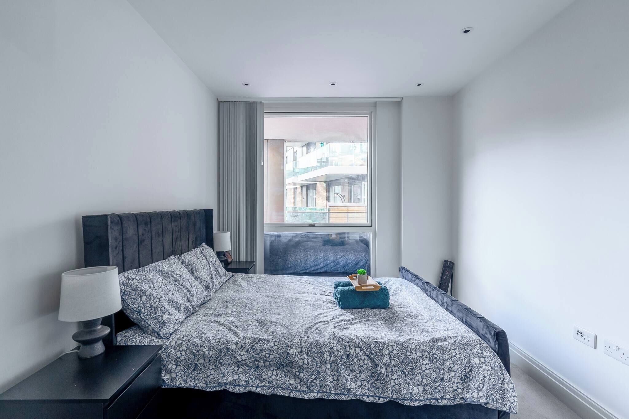 A modern bedroom with a large bed, a bedside table with a lamp, a window with a view of buildings, and a small plant on the bed.