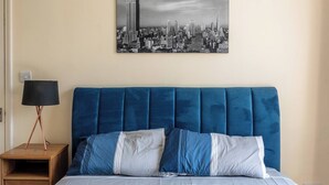 A bed with a blue upholstered headboard, a cityscape painting above it, and a bedside table with a lamp. - Elegant 3BR Victorian Retreat, Free Parking (Kingston Upon Thames)