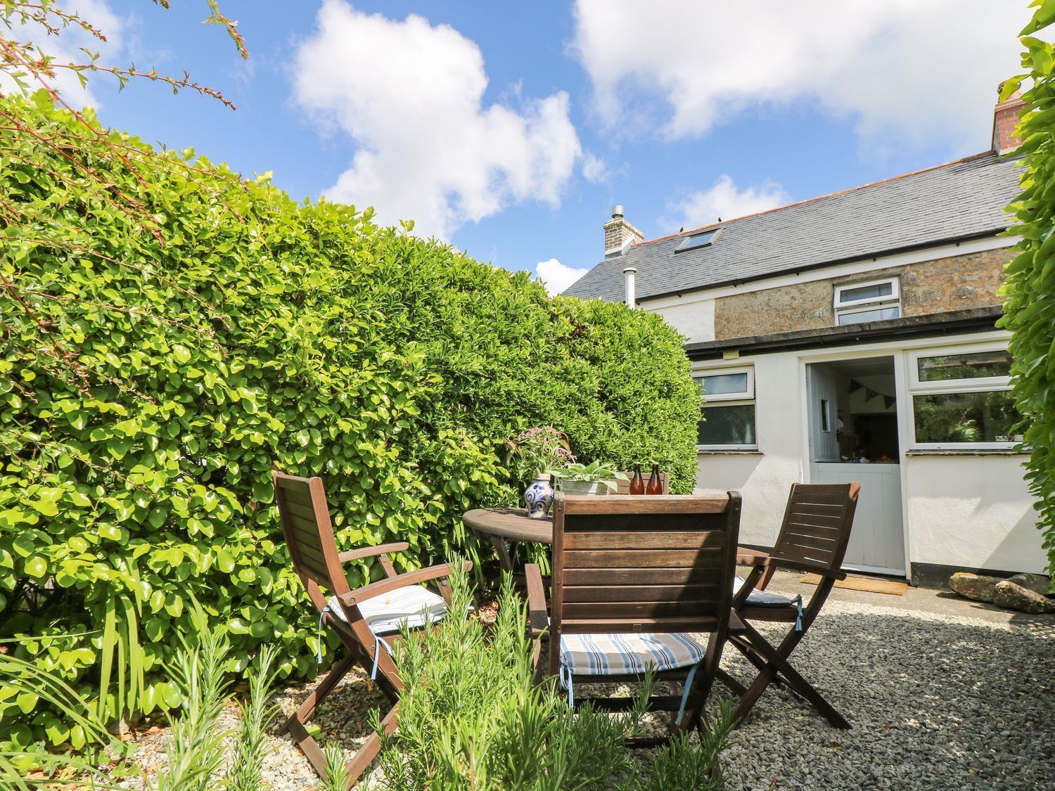 A patio with two wooden chairs and a table, surrounded by a lush green hedge.