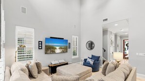Een woonkamer met een beige bank, een poef en een blauwe fauteuil. Er is een flatscreen televisie aan de muur bevestigd, een kleine tafel met een lamp en een raam met jaloezieën.