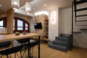 A modern kitchen with a wooden dining table, black bar stools, a TV mounted on a stone wall, and a spiral staircase. - Hanim Boutique | Maisonette with hot tub Old Town, Apartment (Chania)