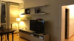 Uma sala de estar com televisão, estantes de livros, uma mesa e um abajur.