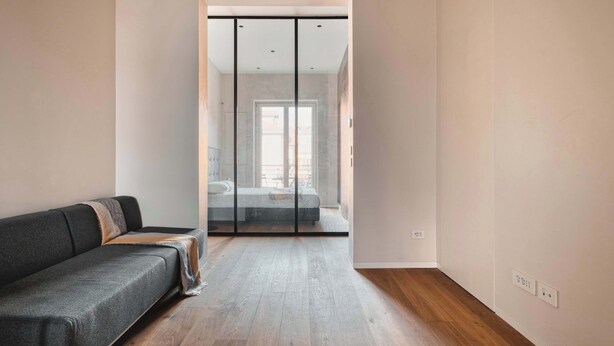 A modern room with a grey sofa, wooden flooring, and a sliding glass door leading to a bedroom with a bed and a window. - w Modern 1BR w Balcony in Porta Venezia (Milan)