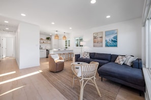A modern living room with a dark blue sofa, a wicker chair, and a small table. There is a kitchen in the background with a white countertop and pendant lights. - Carlsbad Bungalow Large Backyard (Carlsbad)