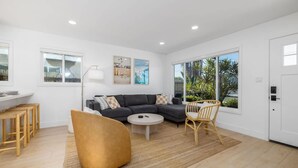 Uma sala de estar moderna com um sofá modular cinza escuro, uma mesa de centro redonda e um banquinho amarelo. Há duas cadeiras de vime, um abajur de chão branco e obras de arte nas paredes.