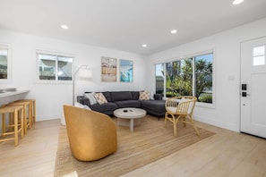 A modern living room with a dark grey sectional sofa, a round coffee table, and a yellow pouf. There are two wicker chairs, a white floor lamp, and artwork on the walls. - Carlsbad Bungalow Large Backyard (Carlsbad)