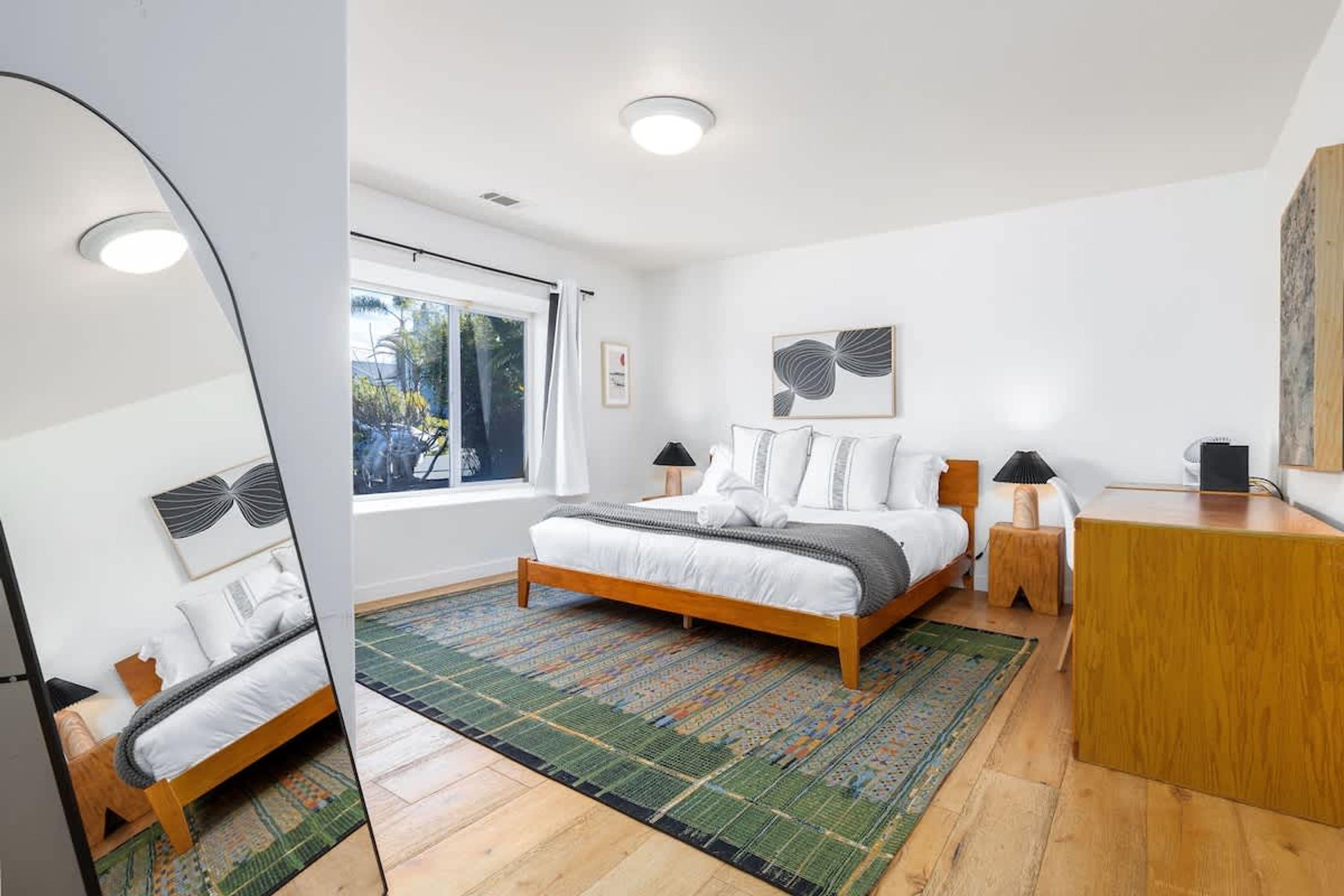 A bedroom with a bed, bedside tables, a desk, a window, and a mirror.