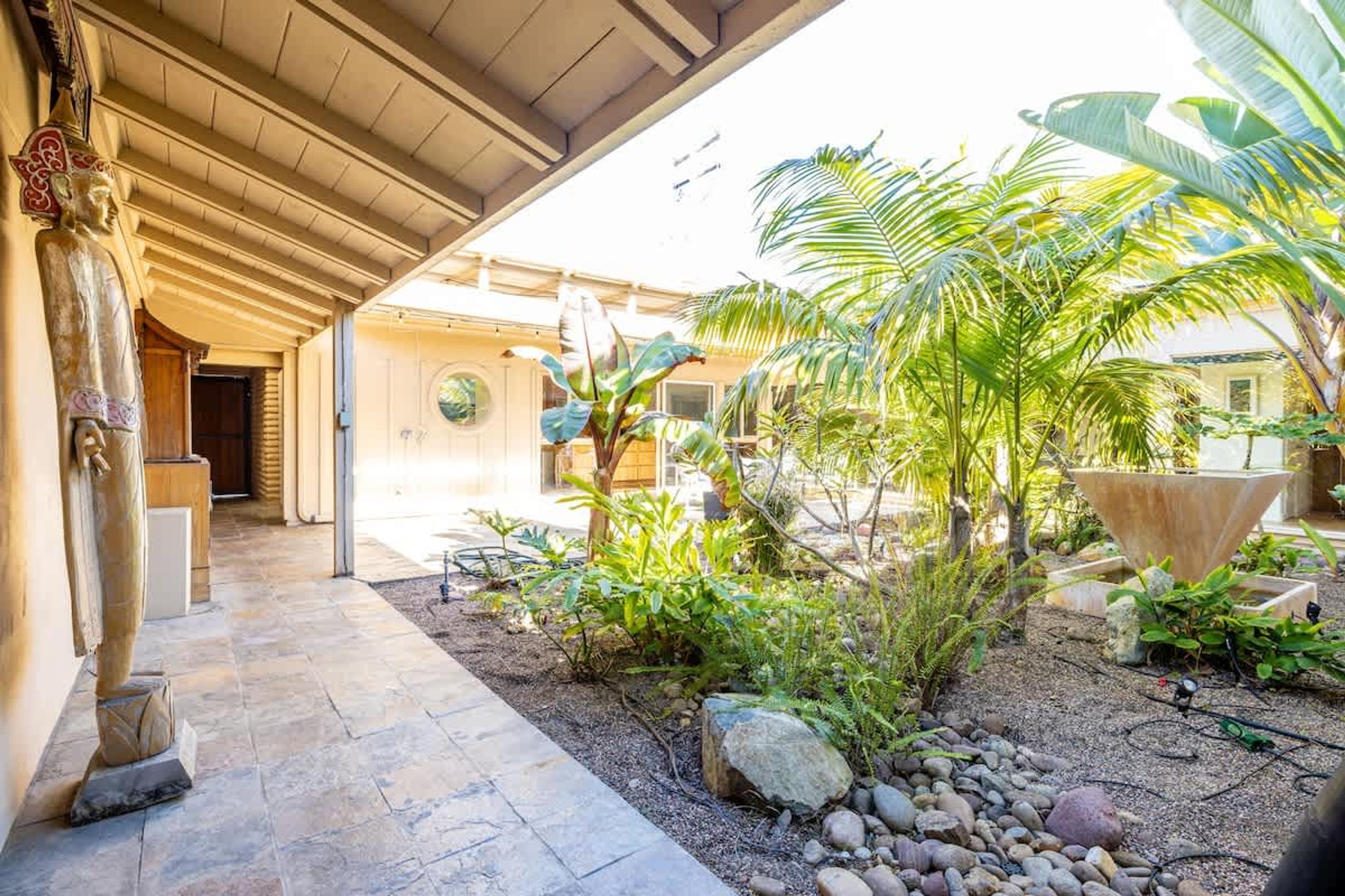 A covered outdoor area with a Buddha statue, palm trees, and a garden with rocks and plants.