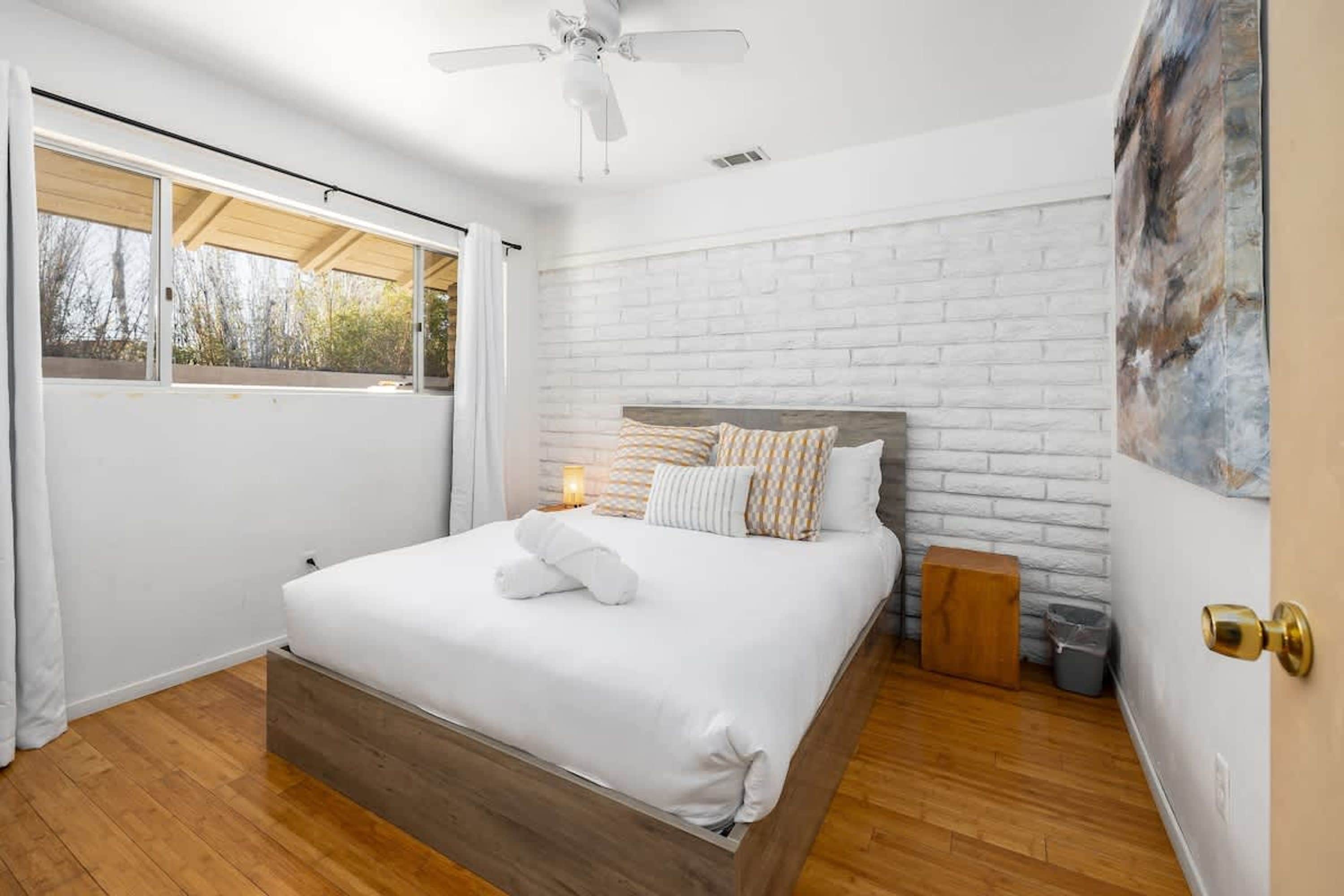 Une chambre avec un lit, un ventilateur de plafond, une fenêtre et un mur en briques blanches.